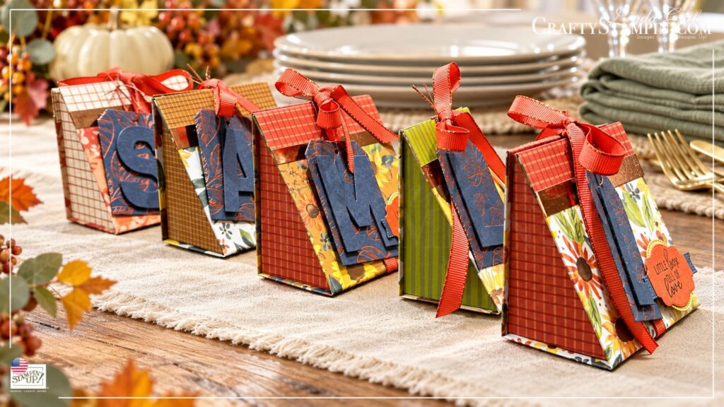 Five boxes side view with stacked plates and napkins Five Thanksgiving mini tea boxes with personalized monogram tags, autumn papers, and coral ribbon beside stacked plates and sage napkins.