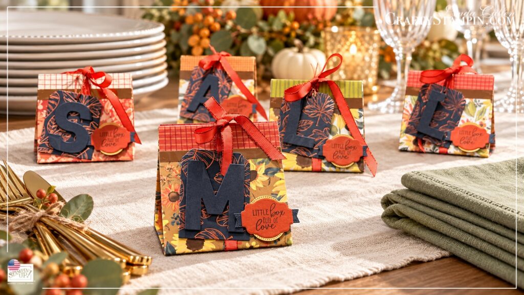 Five boxes with stacked plates and napkins Five Thanksgiving mini tea boxes with personalized monogram tags, autumn papers, and coral ribbon beside stacked plates and sage napkins.